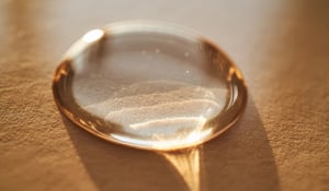 Close-up image of a single water droplet resting on a textured, light-brown surface, illuminated by warm lighting that casts soft reflections and shadows around the droplet.