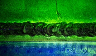 Fluorescent penetrant inspection image showing a weld with visible indications and cracking, illuminated under UV light with a measurement scale below.